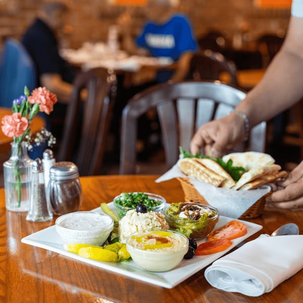 A server places a basket of pita bread next to a platter of Persian appetizers on a wooden table.