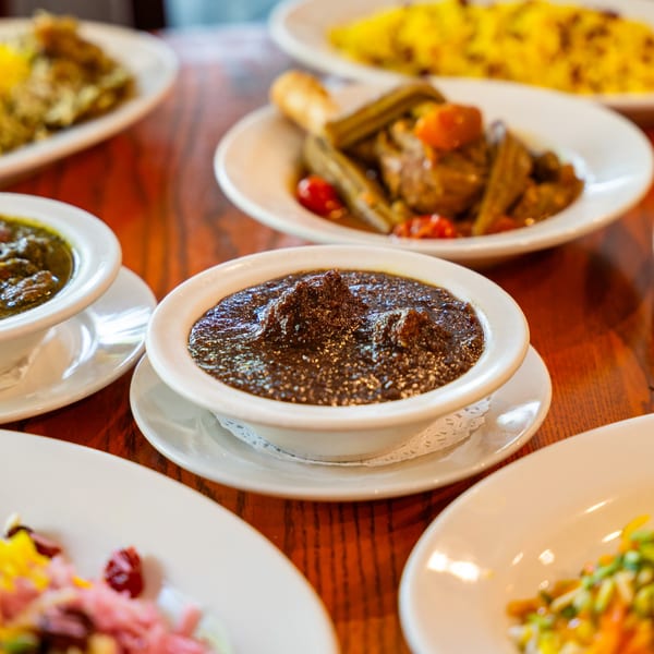 A close-up of a dark Persian stew in a white bowl, surrounded by other dishes on a wooden table.