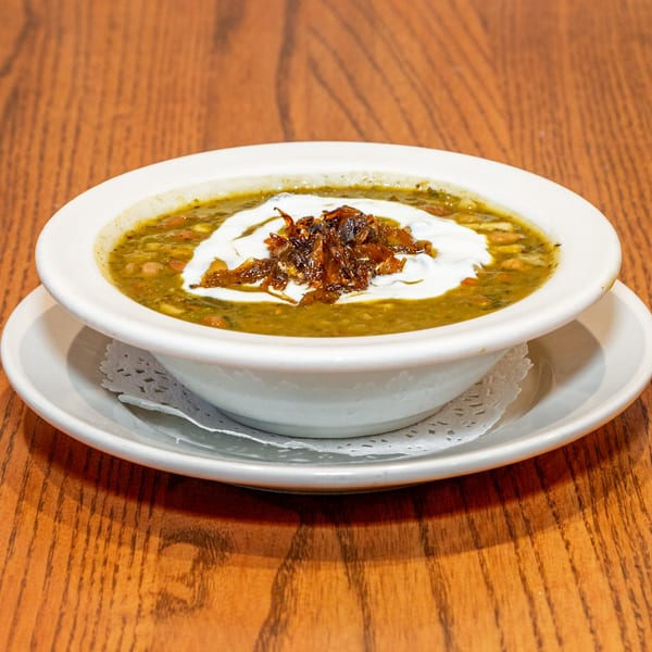 A bowl of Persian Ash Reshteh soup, topped with white cream and crispy fried onions, on a wooden table.